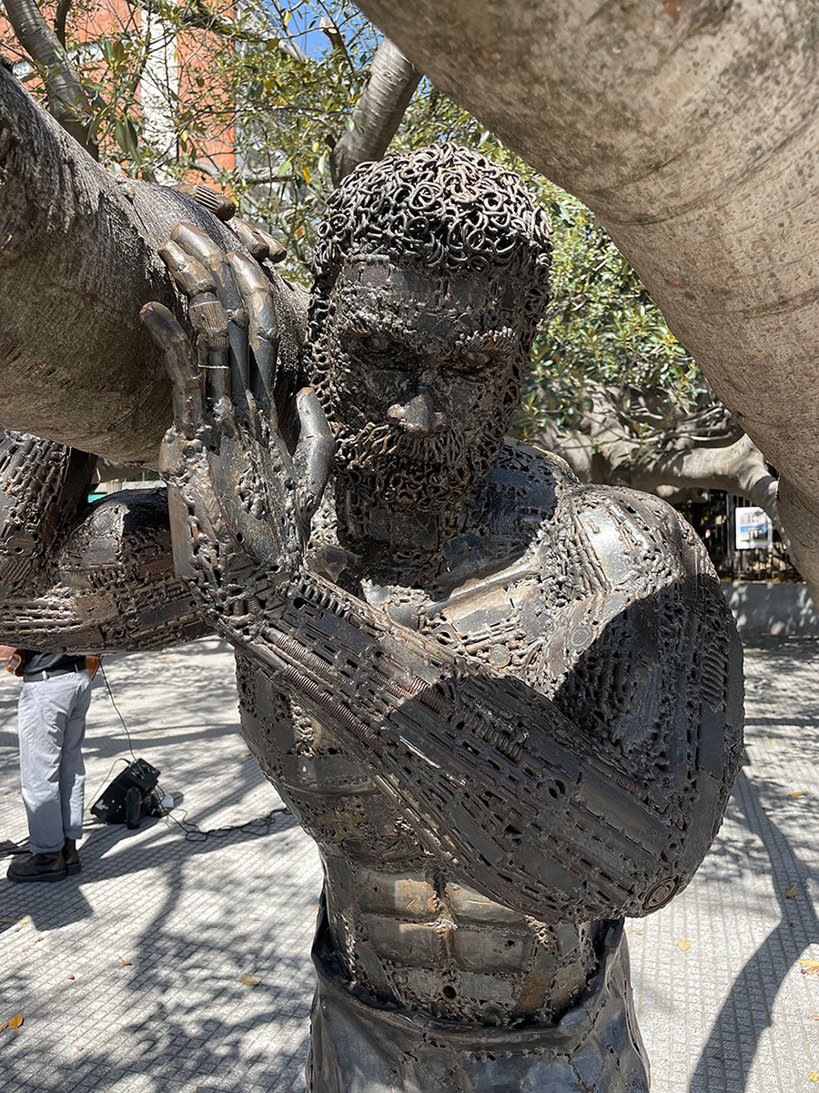 Metallskulptur eines muskulösen Mannes mit Bart, der einen großen zylindrischen Gegenstand auf der Schulter trägt, umgeben von Bäumen und Sonnenlicht.