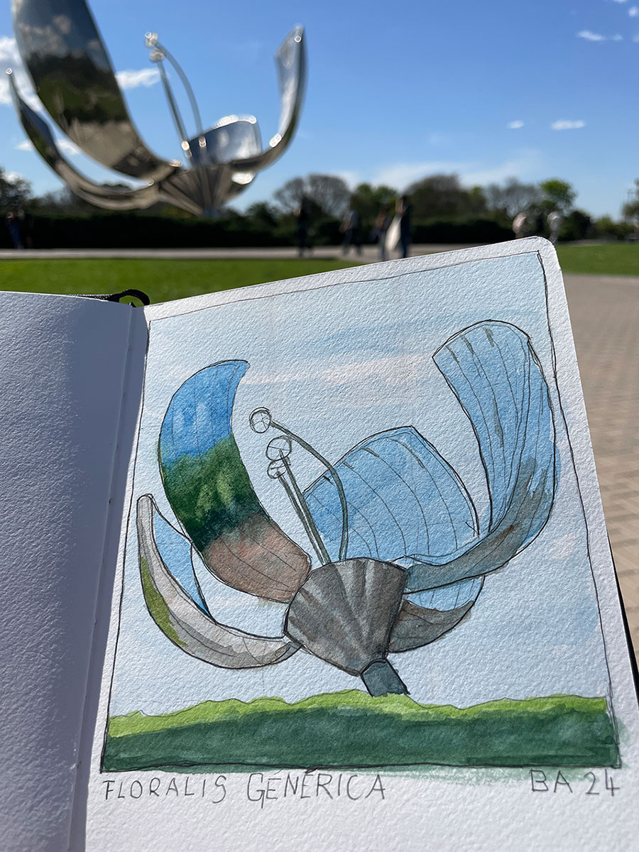 Eine Hand hält eine Aquarellskizze der Skulptur „Floralis Genérica“, die zu dem großen Metallblumen-Denkmal im Hintergrund passt – an einem sonnigen Tag in einem Park.