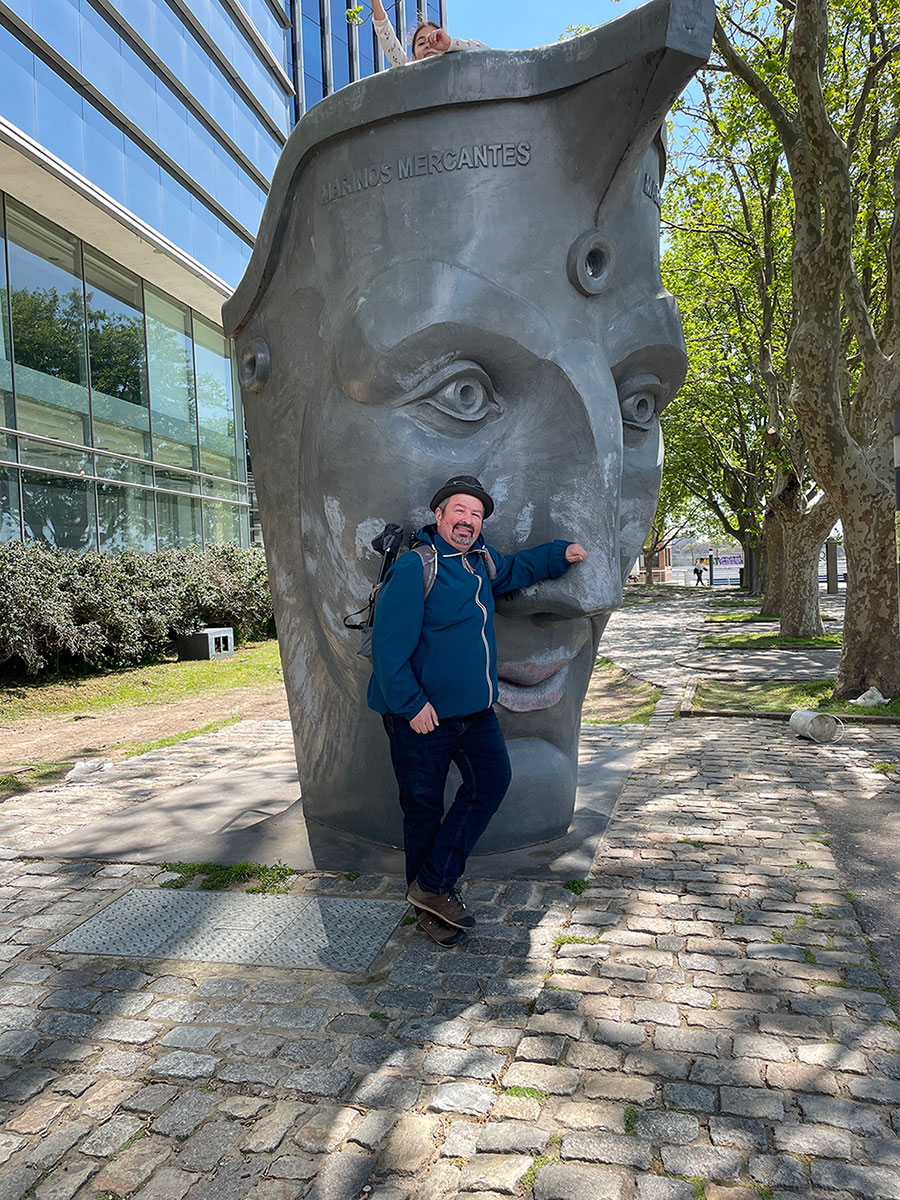 Ein lächelnder Mann in einer blauen Jacke posiert im Freien auf einem Kopfsteinpflasterweg neben einem großen Skulpturenkopf, im Hintergrund sind Bäume und ein modernes Glasgebäude zu sehen.